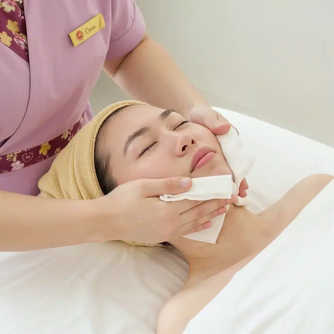 A person with a towel wrapped around their head receives an acne facial from a therapist in a relaxing spa setting.