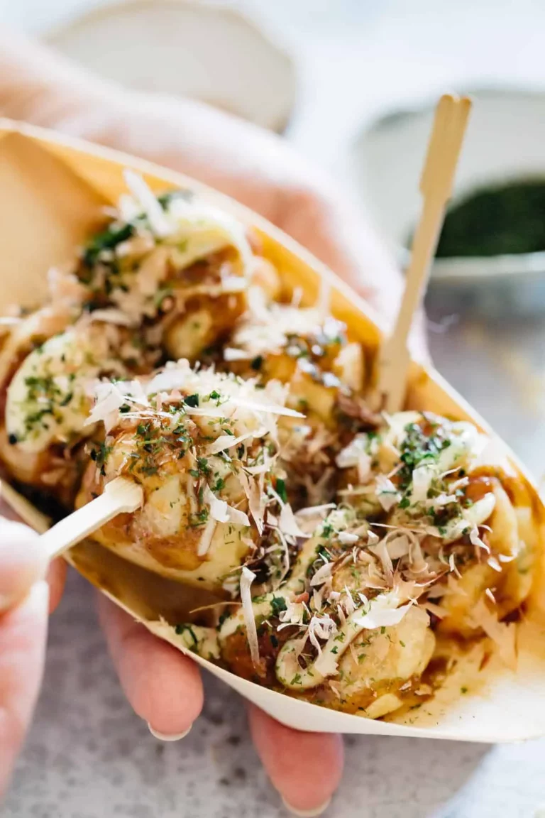 A hand holds a tray of takoyaki, Japanese octopus balls, topped with sauce, mayonnaise, seaweed, and bonito flakes, with wooden skewers for eating. In the background, a chilled bottle of ramune adds a refreshing touch to this classic street food scene.