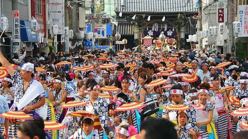 Large crowd participates in a Japanese street festival, with many people wearing traditional clothing and colourful hats. Banners and decorations line the busy street, while vendors offer treats like ramune to refresh festival-goers.