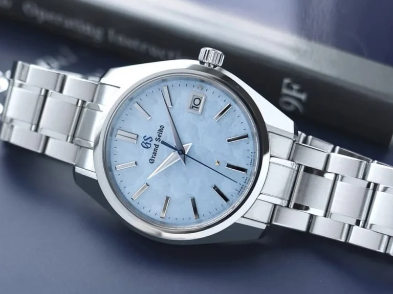 A stainless steel Grand Seiko wristwatch with a light blue textured dial, date window at 3 o’clock, and silver hour markers is elegantly placed on a dark blue surface near a book.