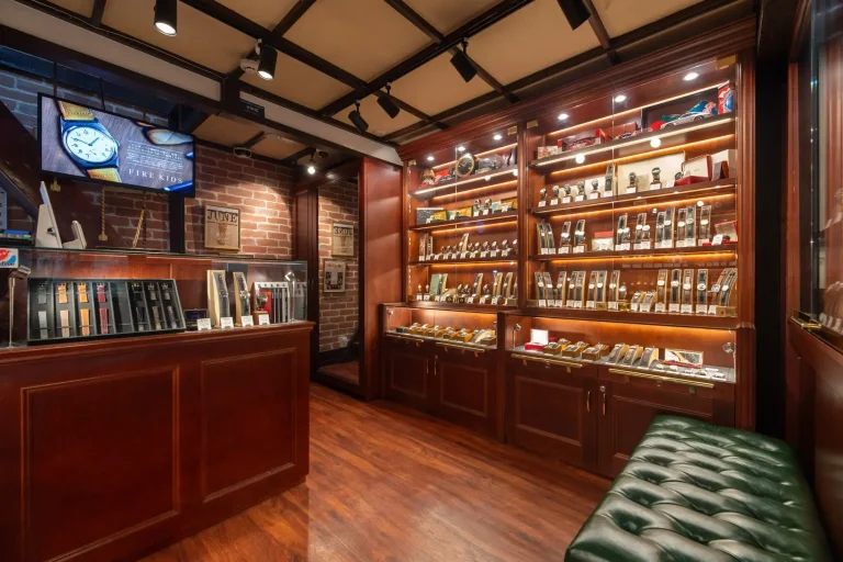 A cigar shop interior with wooden shelves displaying cigars, a counter with books, brick feature wall, clock, and a green leather bench on a polished wooden floor. A Grand Seiko watch rests elegantly atop the counter, adding a touch of refined luxury to the inviting space.