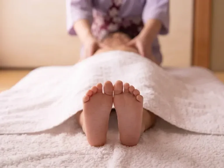 Person lying face up under a white blanket with bare feet visible, enjoying a relaxing massage from someone wearing a light dressing gown.