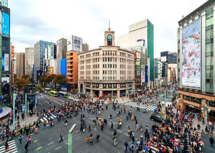 A busy city crossroads with many people crossing, surrounded by tall buildings—including a central building with a clock tower reminiscent of the precision and elegance found in a Grand Seiko timepiece.