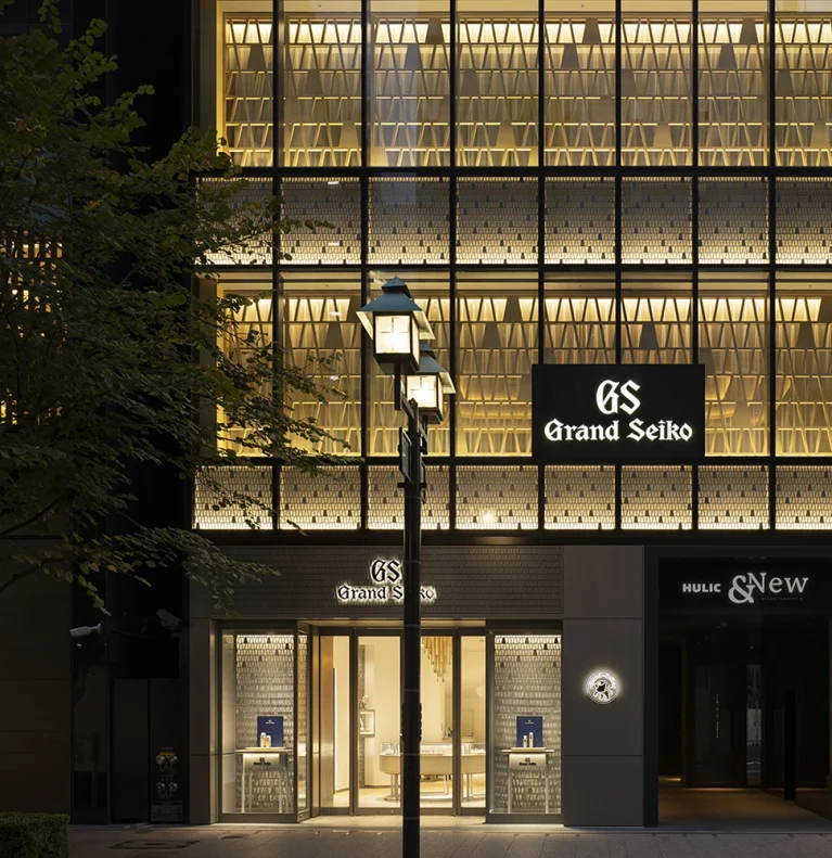 Exterior view of a Grand Seiko shop at night, with illuminated Grand Seiko signage and warm lighting visible through large windows.
