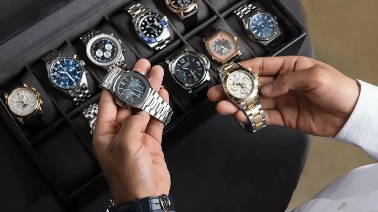 A person holds two luxury wristwatches, including a stunning Grand Seiko, above an open watch case displaying an assortment of watches with various designs and colours.