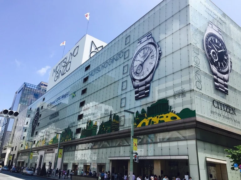 Large department store with a gleaming glass exterior, featuring two giant Grand Seiko watch advertisements and crowds of people at the entrance on a sunny day.