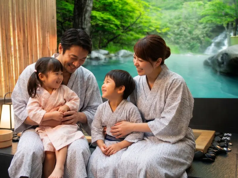 A family of four in matching yukata sit together and smile by an outdoor hot spring, with lush greenery and water in the background, enjoying a relaxing children's spa experience.