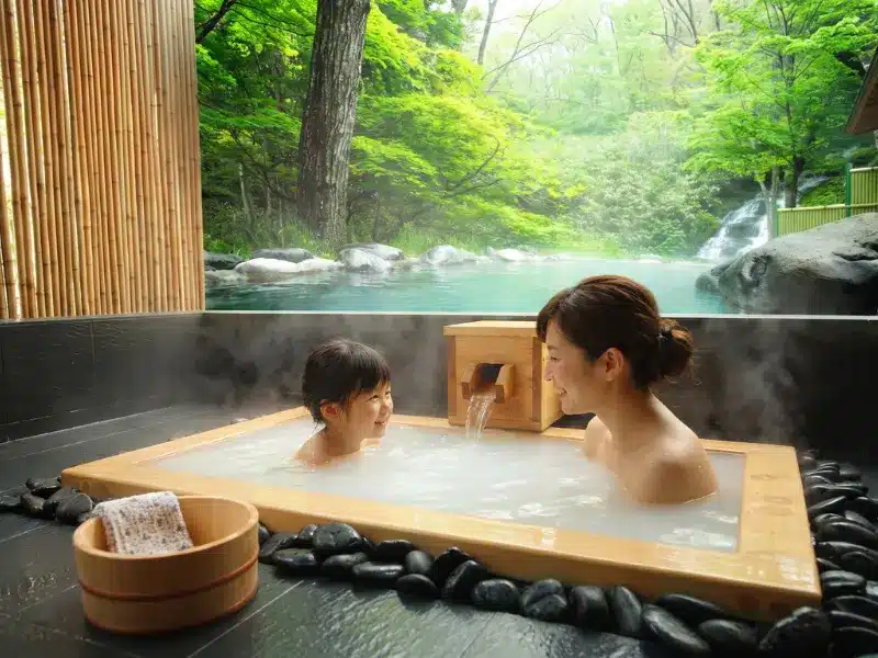A woman and child enjoy a hot spring bath indoors at this children's spa, surrounded by stones, with lush green trees and a stream visible through an open wall in the background.