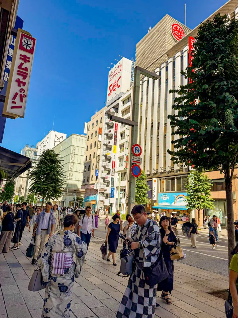 Crowds walk along a busy city street lined with trees and tall buildings, with people in traditional and modern clothing—some glancing at their Grand Seiko watches—under a clear blue sky.