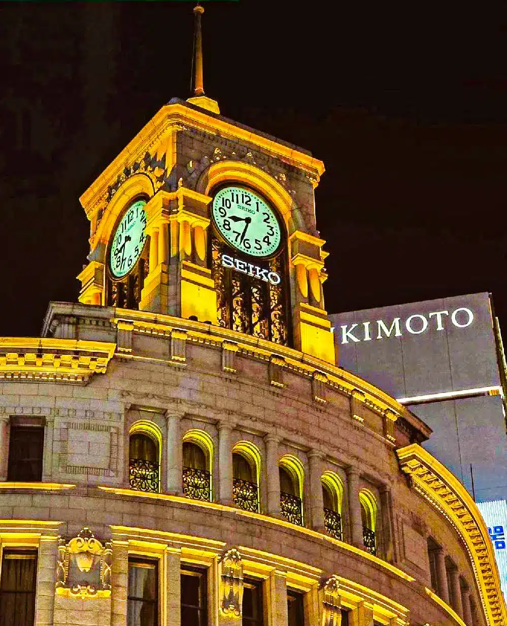 A clock tower illuminated at night, displaying the Seiko brand above its clock face—a nod to the precision of Grand Seiko—whilst a nearby building shows the name Kimoto.