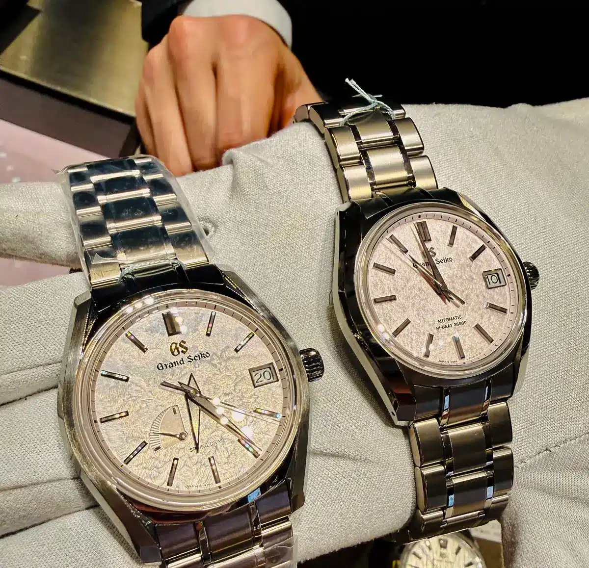 Two elegant Grand Seiko stainless steel wristwatches with textured light-coloured dials are displayed on a padded stand, with a person’s hand visible in the background.