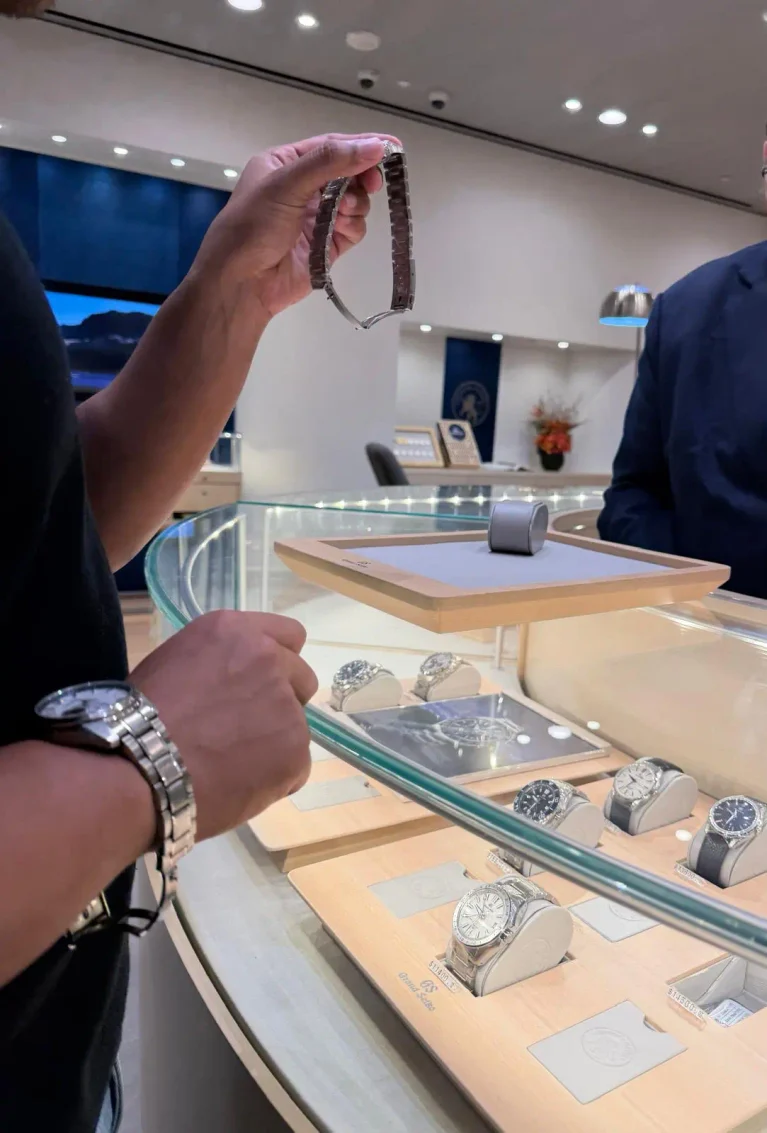 A person at a jewellery shop counter holds a Grand Seiko wristwatch while several luxury watches are displayed in a glass case.