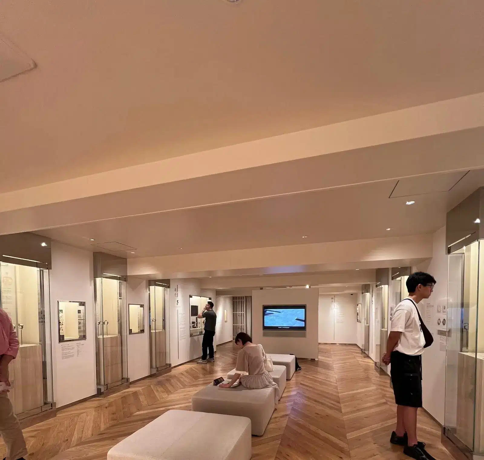 People observe exhibits and displays inside a well-lit museum room with wooden floors, glass cases, and cushioned benches, where a special showcase features the craftsmanship of Grand Seiko timepieces.