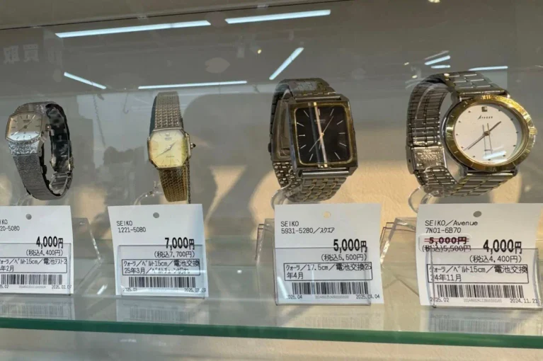 Four Seiko wristwatches, including a striking Grand Seiko model, are displayed on a glass shelf, each with a price tag in yen beneath them, inside a shop setting with overhead lighting reflected on the glass.