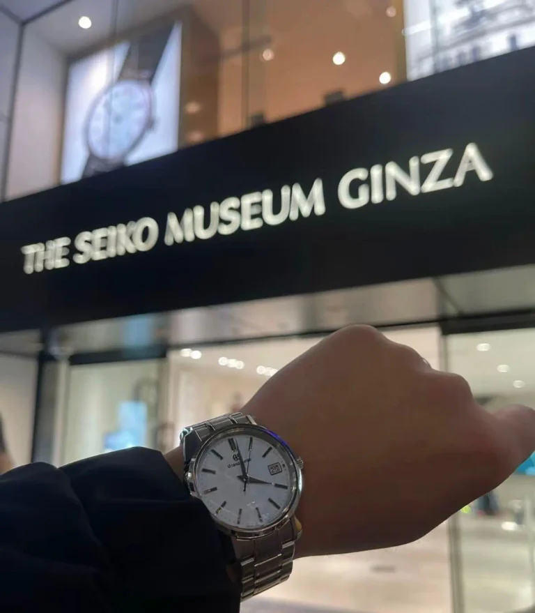 A person shows a silver Grand Seiko wristwatch in front of the entrance to The Seiko Museum Ginza, with the museum sign visible in the background.