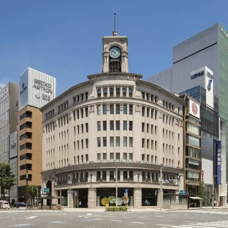 A large historic building with a central clock tower stands at a street junction, surrounded by modern high-rise buildings under a clear blue sky—a timeless scene where tradition meets innovation, much like the elegance embodied by Grand Seiko.