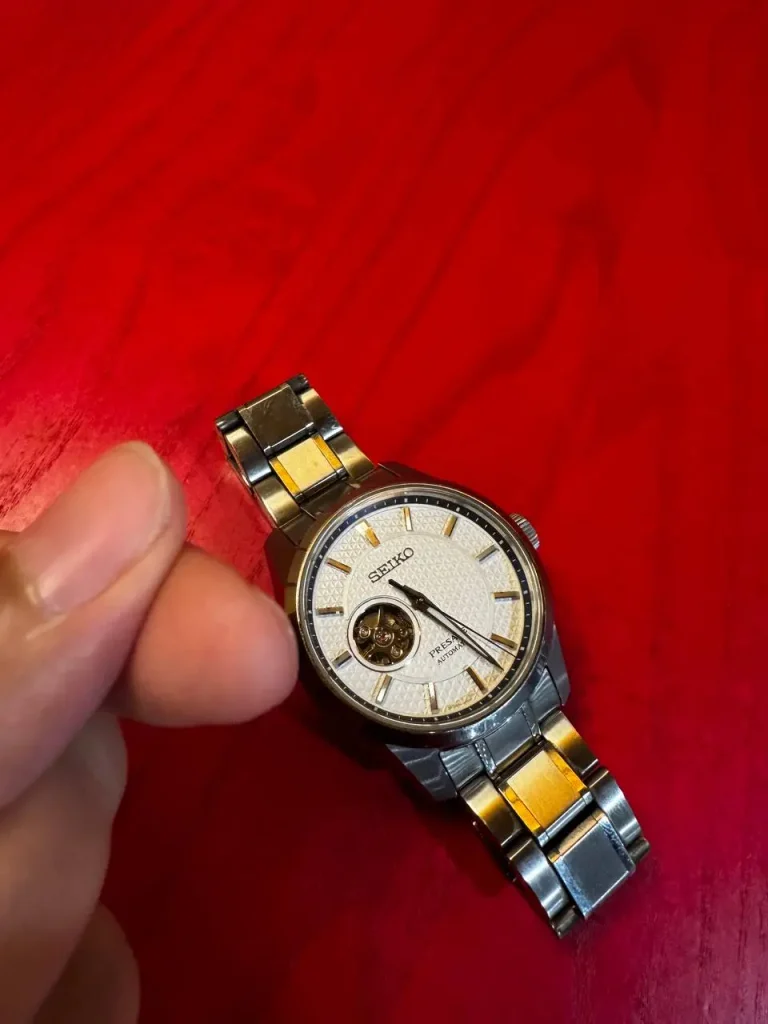 A hand is pointing near a Grand Seiko wristwatch with a gold and silver metal strap on a red wooden surface.