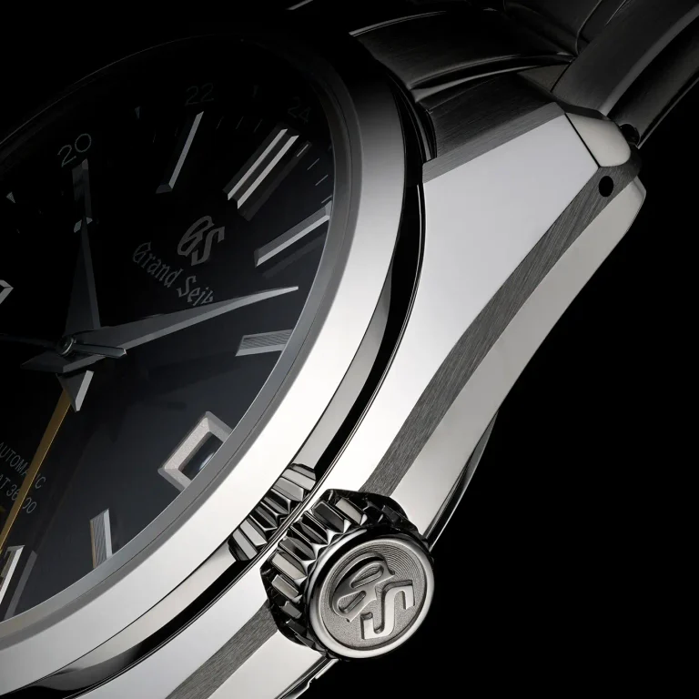 Close-up of a Grand Seiko wristwatch, highlighting the crown with the GS logo, the meticulously crafted stainless steel case, and part of the elegant black dial with crisp white hour markers and hands—a true testament to Grand Seiko’s precision and refinement.