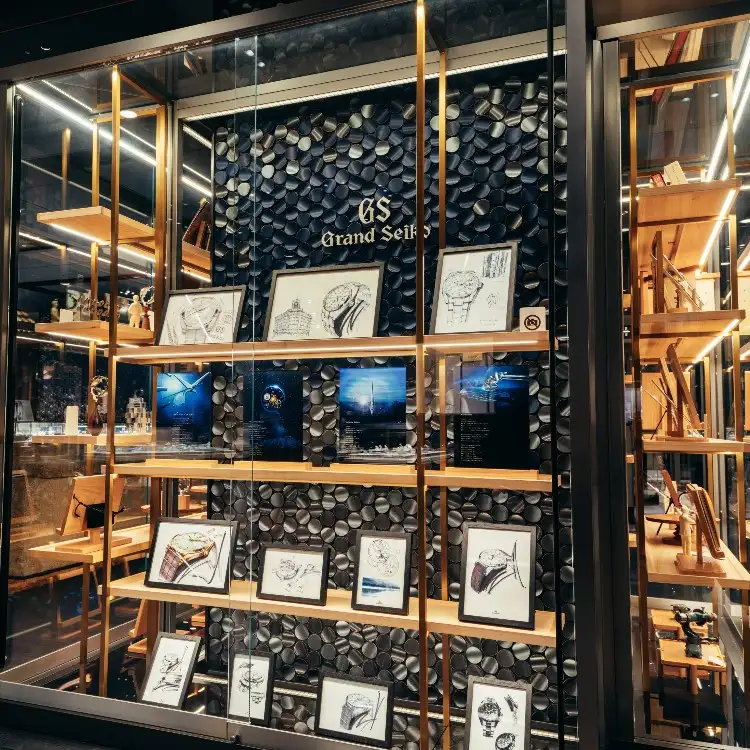 Shopfront display for Grand Seiko featuring framed Grand Seiko watch sketches, photographs, and models on wooden shelves against a decorative black textured wall.