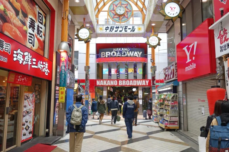People walk through a covered shopping arcade with shops and signs, including a Grand Seiko boutique, leading to the entrance of Nakano Broadway in Tokyo, Japan.