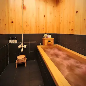 A traditional Japanese wooden soaking tub filled with water sits in a black-tiled room with light wood walls, offering the perfect retreat for relaxation. Bottles and a wooden stool are placed beside the tub, creating an inviting spa deal atmosphere that promises tranquillity and rejuvenation.