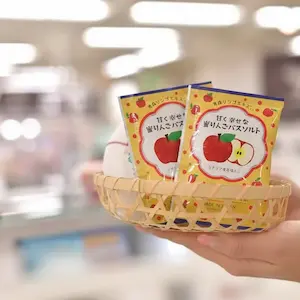 A hand holds a small wicker basket containing two apple-themed packaged products with Japanese text, set in a brightly lit shop that offers an exclusive Spa Deal.