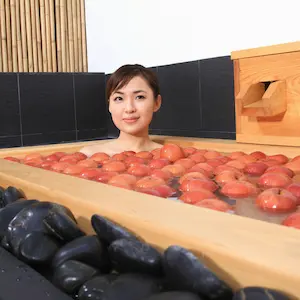 Woman enjoying a relaxing spa offer, sitting in a wooden tub filled with water and floating red apples, with black stones placed on the edge of the bath.